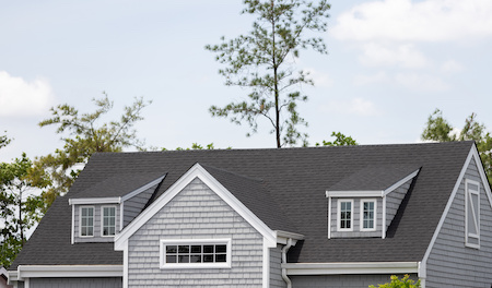 Roof Shingles With Garret House On Top Of The House Among A Lot Of Trees. Dark Asphalt Tiles On The Roof With White Rain Gutter And Down Pipe.
