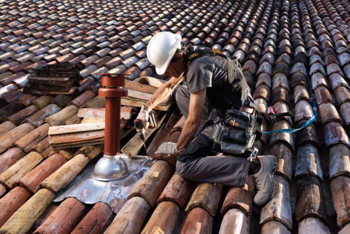 Professional Roofer Working On A Residential Clay Tile Roof, Installing A Coaxial Boiler Flue With Safety Harness. Home Improvement, Construction Safety, And Roofing Services Concept.